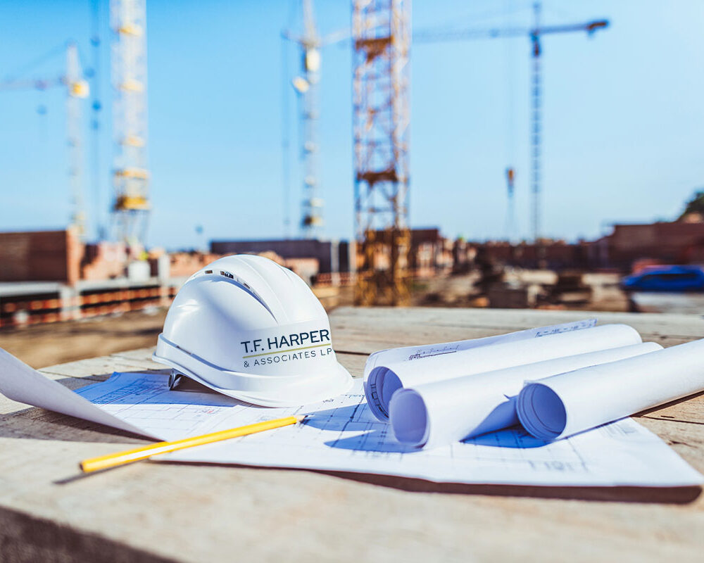 Building plans and a hardhat are on a construction job site with cranes in the background.