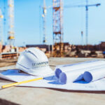 Building plans and a hardhat are on a construction job site with cranes in the background.