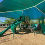 A green shade structure is protecting an outdoor playground from the sun.