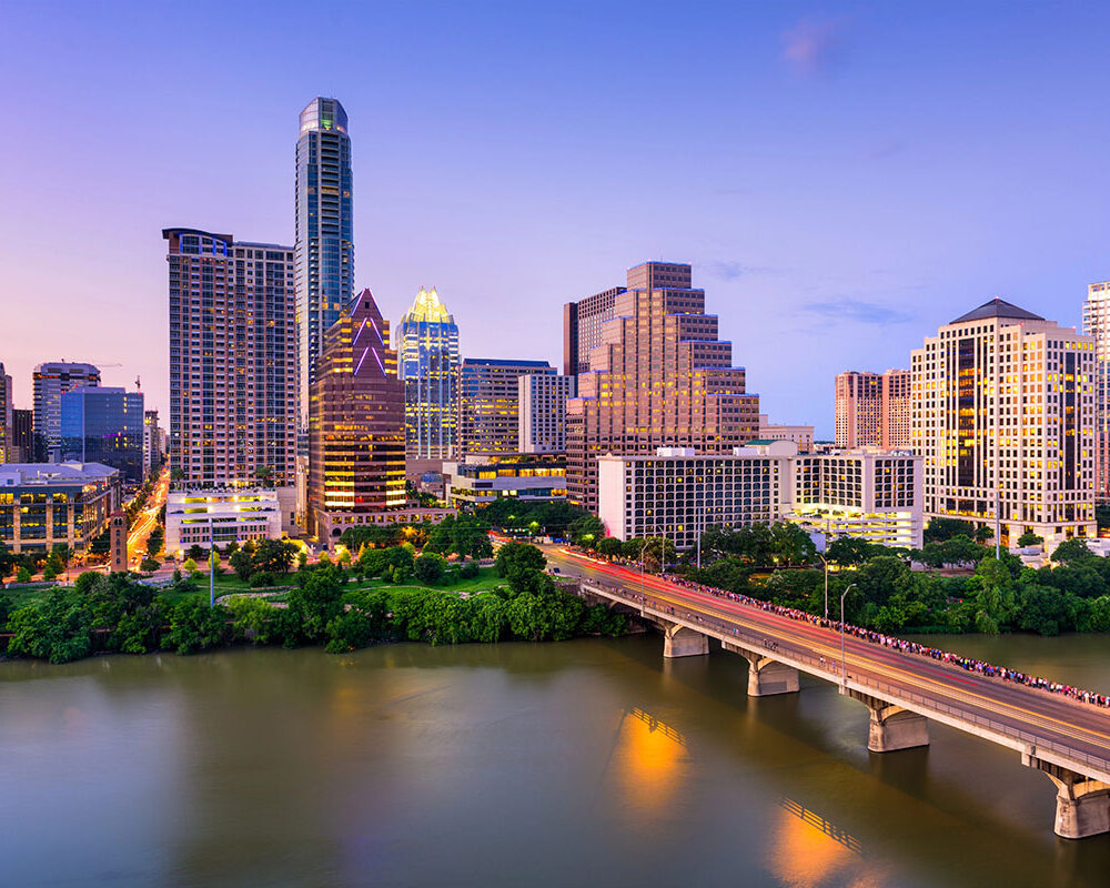 Austin skyline at dusk, featuring a mix of modern skyscrapers, the Colorado River in the foreground, and a bridge with a crowd of pedestrians crossing. The sky displays soft hues of blue and purple.