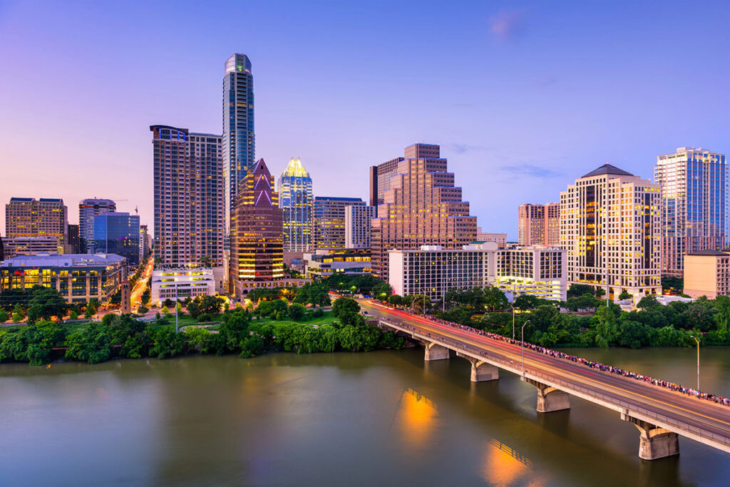Austin skyline at dusk, featuring a mix of modern skyscrapers, the Colorado River in the foreground, and a bridge with a crowd of pedestrians crossing. The sky displays soft hues of blue and purple.