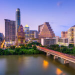 Austin skyline at dusk, featuring a mix of modern skyscrapers, the Colorado River in the foreground, and a bridge with a crowd of pedestrians crossing. The sky displays soft hues of blue and purple.