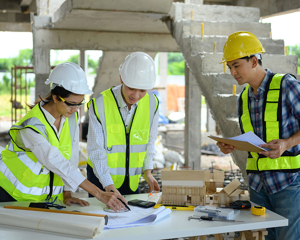 Three construction professionals, wearing hard hats and high-visibility vests, collaborate over a table filled with blueprints, a calculator, and construction tools at a building site. One person points at the papers while another holds a clipboard, and a miniature building model is displayed on the table.