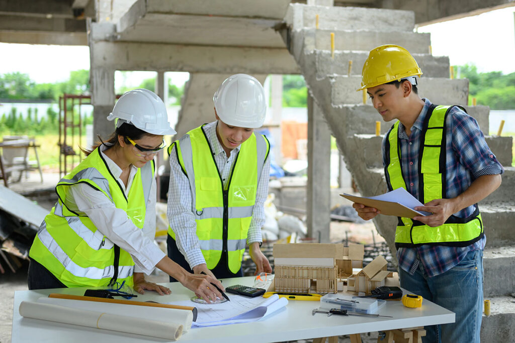Three construction professionals, wearing hard hats and high-visibility vests, collaborate over a table filled with blueprints, a calculator, and construction tools at a building site. One person points at the papers while another holds a clipboard, and a miniature building model is displayed on the table.