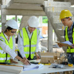 Three construction professionals, wearing hard hats and high-visibility vests, collaborate over a table filled with blueprints, a calculator, and construction tools at a building site. One person points at the papers while another holds a clipboard, and a miniature building model is displayed on the table.