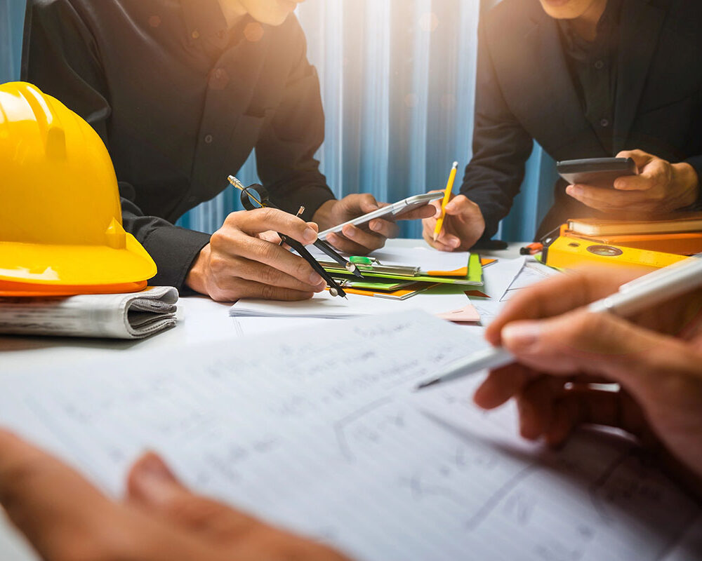 A group of professionals in black shirts collaborating at a table, surrounded by construction tools, papers, and a yellow hard hat. One person is writing with a pen, while others are using smartphones and handling colorful stationery.