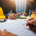 A group of professionals in black shirts collaborating at a table, surrounded by construction tools, papers, and a yellow hard hat. One person is writing with a pen, while others are using smartphones and handling colorful stationery.