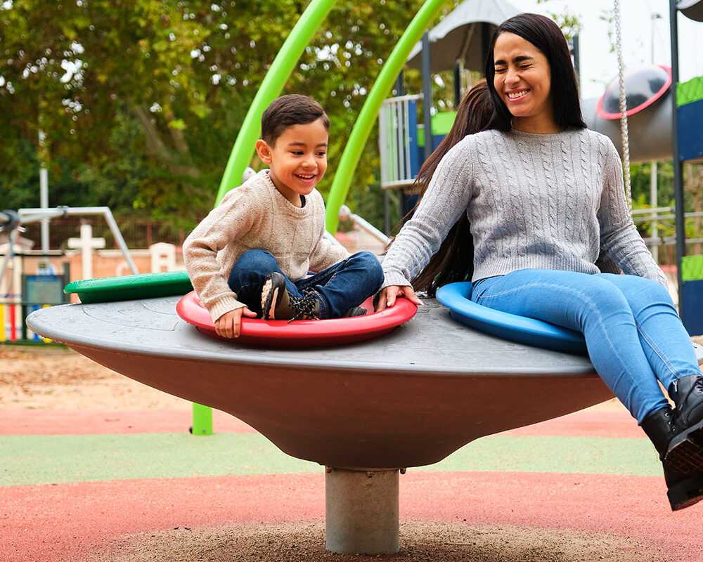 A woman and a young boy are enjoying a spinning playground ride, sitting on colorful circular seats. The boy smiles widely, while the woman laughs with her eyes closed, both clearly having fun in a vibrant outdoor park setting.