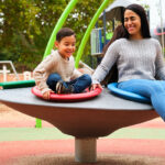 A woman and a young boy are enjoying a spinning playground ride, sitting on colorful circular seats. The boy smiles widely, while the woman laughs with her eyes closed, both clearly having fun in a vibrant outdoor park setting.