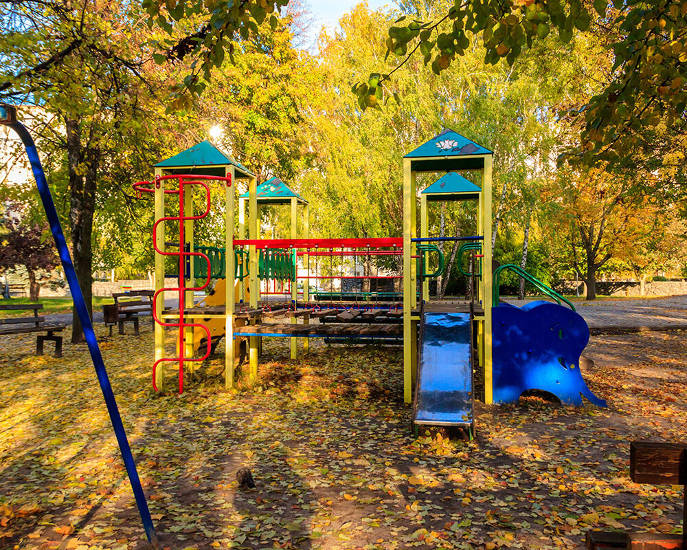 A colorful playground with a climbing structure, slide, and swings, surrounded by trees displaying autumn leaves and benches in a park setting.