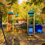 A colorful playground with a climbing structure, slide, and swings, surrounded by trees displaying autumn leaves and benches in a park setting.