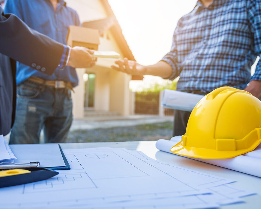 Two individuals are exchanging documents and a small box outdoors, with architectural plans and a yellow hard hat visible on a table in the foreground.
