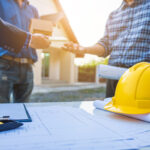 Two individuals are exchanging documents and a small box outdoors, with architectural plans and a yellow hard hat visible on a table in the foreground.