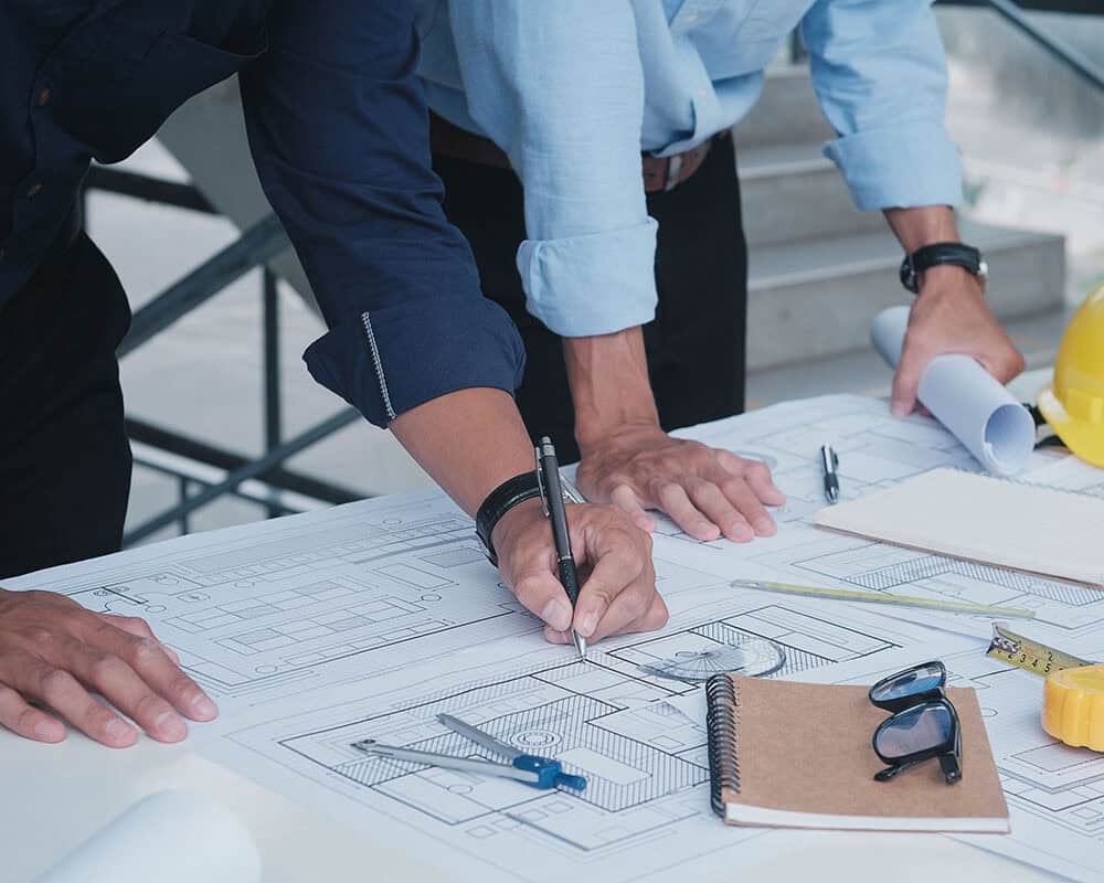 Two professionals are reviewing blueprints on a table. One is using a pen to take notes, while the other stands nearby holding rolled plans. Various drafting tools, a notebook, and safety gear are visible on the table.