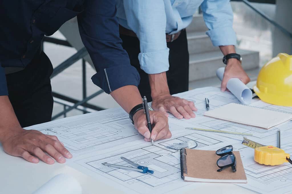Two professionals are reviewing blueprints on a table. One is using a pen to take notes, while the other stands nearby holding rolled plans. Various drafting tools, a notebook, and safety gear are visible on the table.