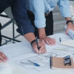 Two professionals are reviewing blueprints on a table. One is using a pen to take notes, while the other stands nearby holding rolled plans. Various drafting tools, a notebook, and safety gear are visible on the table.