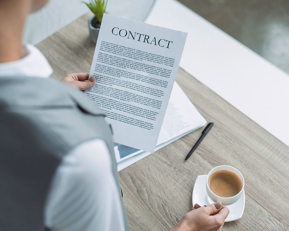 A person holding a contract document while seated at a table with a cup of coffee and a pen nearby.