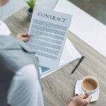 A person holding a contract document while seated at a table with a cup of coffee and a pen nearby.