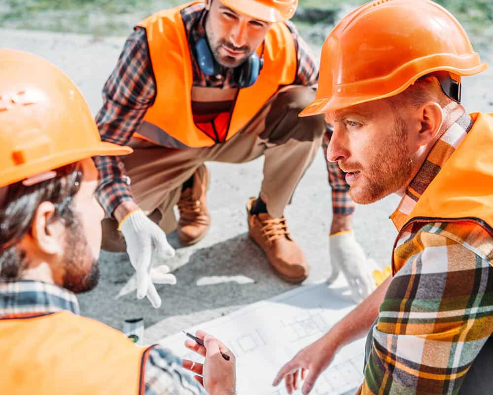 Three construction workers, wearing orange hard hats and vests, are gathered around a blueprint on the ground, discussing plans intently. One worker is pointing to the blueprint, while the others are observing closely.