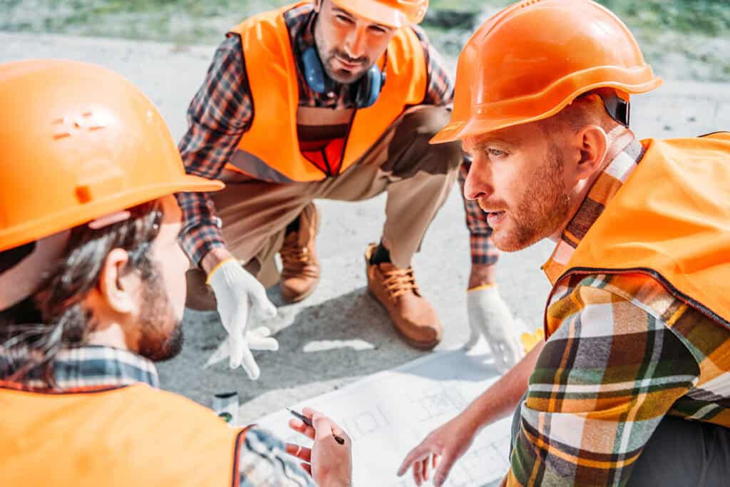 Three construction workers, wearing orange hard hats and vests, are gathered around a blueprint on the ground, discussing plans intently. One worker is pointing to the blueprint, while the others are observing closely.