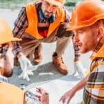Three construction workers, wearing orange hard hats and vests, are gathered around a blueprint on the ground, discussing plans intently. One worker is pointing to the blueprint, while the others are observing closely.