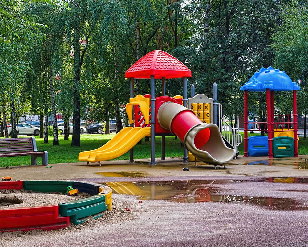 A colorful children's playground featuring a red slide, yellow slide, and a blue play structure, surrounded by trees and a grassy area. A sandbox with colorful borders is visible, along with puddles on the ground.