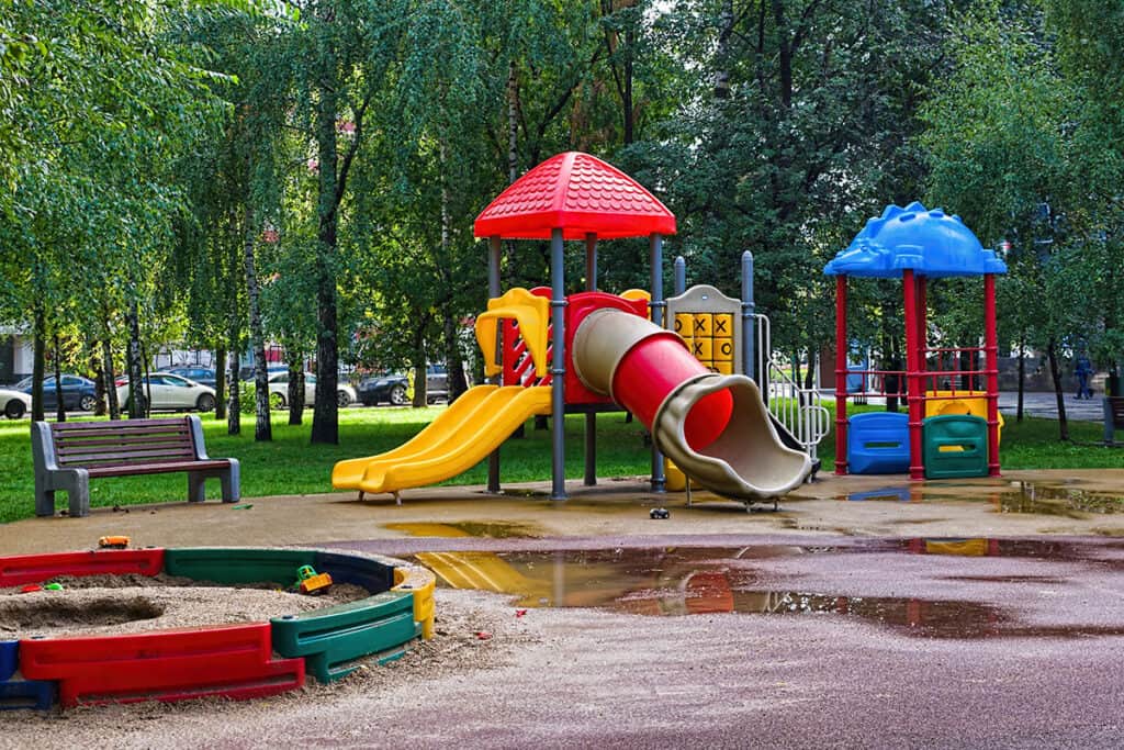 A colorful children's playground featuring a red slide, yellow slide, and a blue play structure, surrounded by trees and a grassy area. A sandbox with colorful borders is visible, along with puddles on the ground.