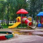 A colorful children's playground featuring a red slide, yellow slide, and a blue play structure, surrounded by trees and a grassy area. A sandbox with colorful borders is visible, along with puddles on the ground.