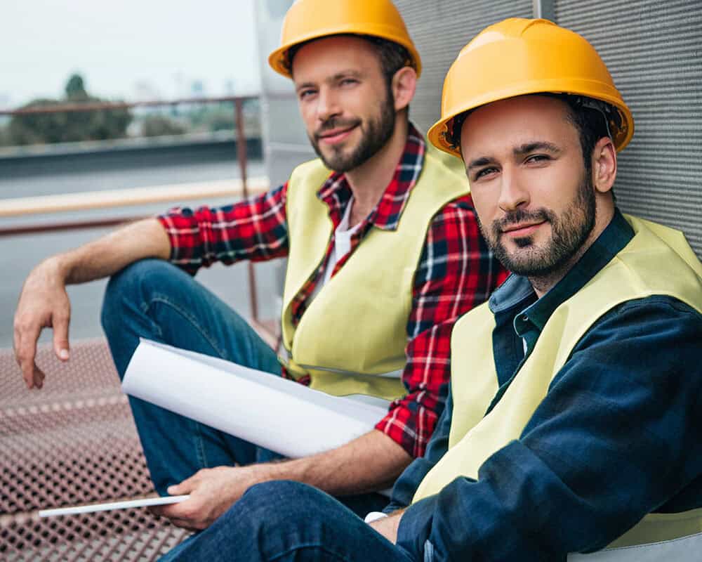 Two smiling construction workers sitting on a rooftop, wearing yellow hard hats and safety vests. One is in a red plaid shirt, while the other wears a blue button-up shirt. They hold blueprints and appear relaxed against a wall.