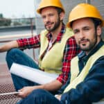 Two smiling construction workers sitting on a rooftop, wearing yellow hard hats and safety vests. One is in a red plaid shirt, while the other wears a blue button-up shirt. They hold blueprints and appear relaxed against a wall.