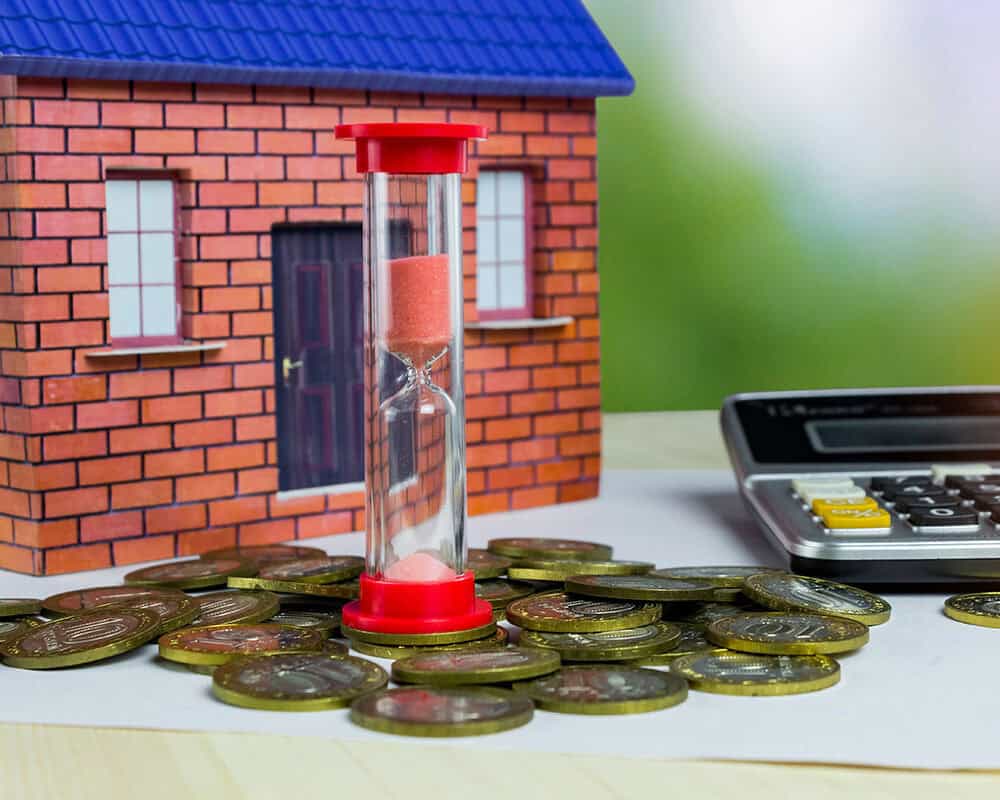 A toy house sits in the background, with an hourglass filled with pink sand positioned in front. Surrounding the hourglass are coins stacked on a white sheet of paper, alongside a calculator.