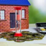 A toy house sits in the background, with an hourglass filled with pink sand positioned in front. Surrounding the hourglass are coins stacked on a white sheet of paper, alongside a calculator.