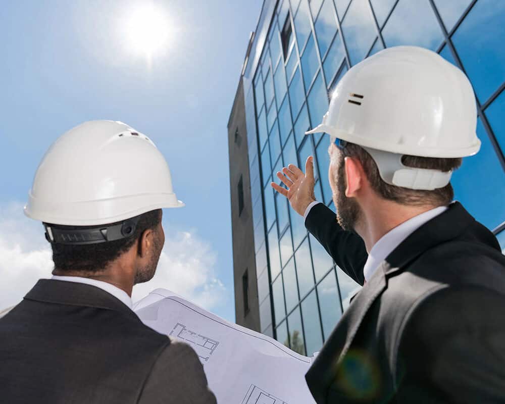 Two professionals in hard hats, one holding blueprints, gaze upward at a modern glass building under a clear blue sky, with the sun shining brightly above.