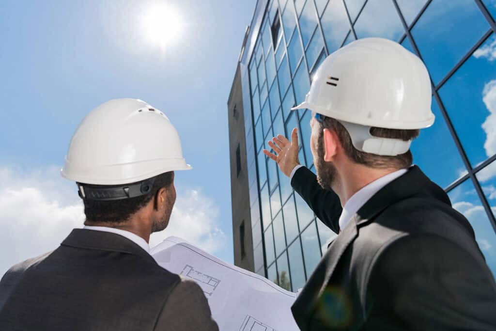 Two professionals in hard hats, one holding blueprints, gaze upward at a modern glass building under a clear blue sky, with the sun shining brightly above.