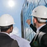 Two professionals in hard hats, one holding blueprints, gaze upward at a modern glass building under a clear blue sky, with the sun shining brightly above.