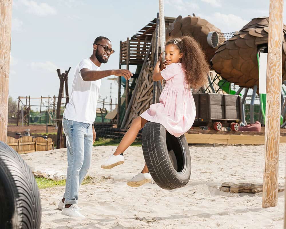 A man in a white shirt assists a smiling girl with curly hair as she swings on a tire at a playground, with play structures in the background under a clear blue sky.
