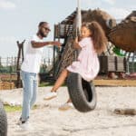 A man in a white shirt assists a smiling girl with curly hair as she swings on a tire at a playground, with play structures in the background under a clear blue sky.