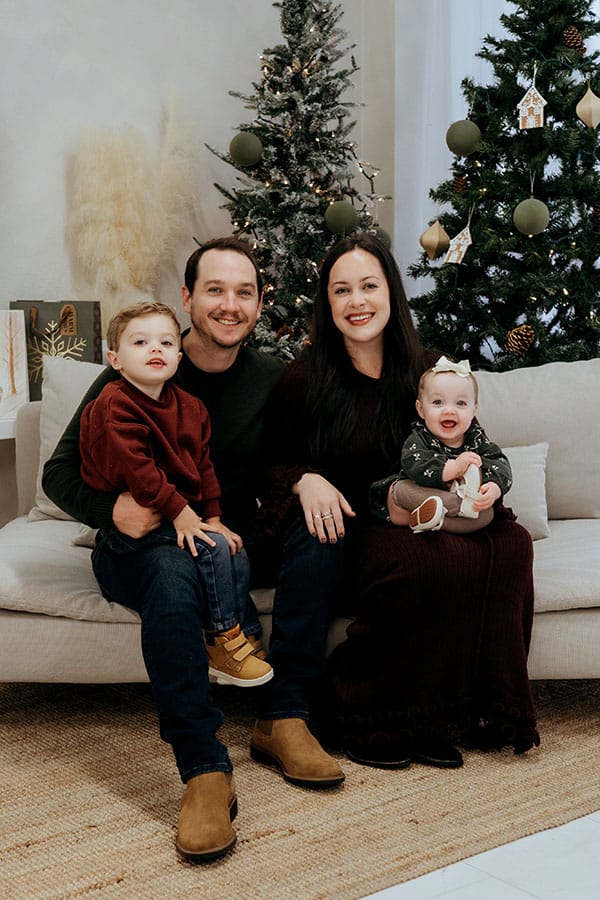 A family of four sits together on a beige couch, smiling at the camera. The father holds a young boy in a red sweater, while the mother cradles a baby girl in a dark outfit. Two decorated Christmas trees stand in the background, adding a festive touch to the cozy scene.