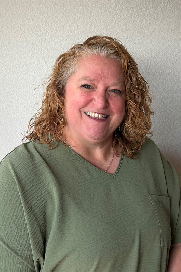 A smiling woman with wavy, shoulder-length blonde hair wears a green v-neck shirt and stands against a light gray wall.