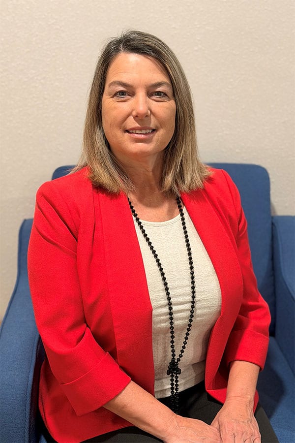 A woman with shoulder-length hair is seated on a blue chair, wearing a red blazer and a white top, adorned with a black necklace. She is smiling at the camera against a light-colored wall.