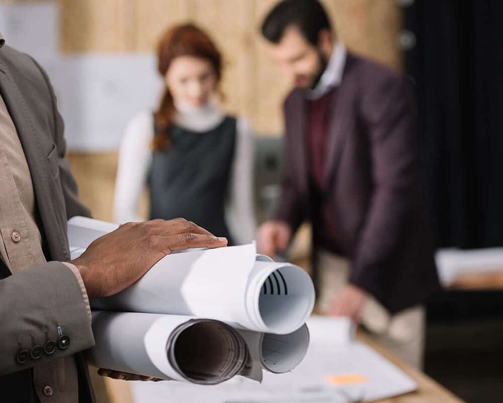 A person in a suit holds a set of blueprints, while two colleagues, a woman in a black dress and a man in a maroon blazer, discuss plans at a table in a modern office setting.
