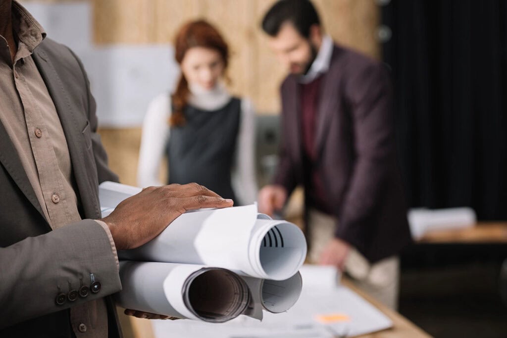 A person in a suit holds a set of blueprints, while two colleagues, a woman in a black dress and a man in a maroon blazer, discuss plans at a table in a modern office setting.