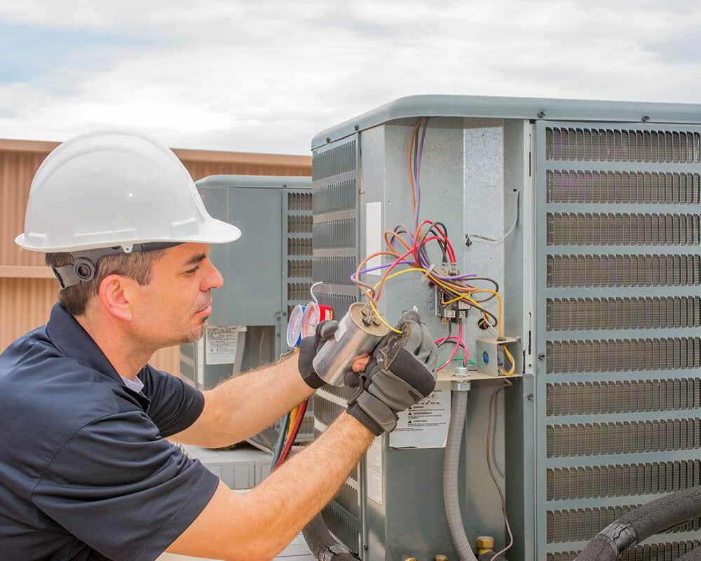 A technician in a hard hat and gloves performing maintenance on an outdoor HVAC unit, inspecting electrical connections and hoses.
