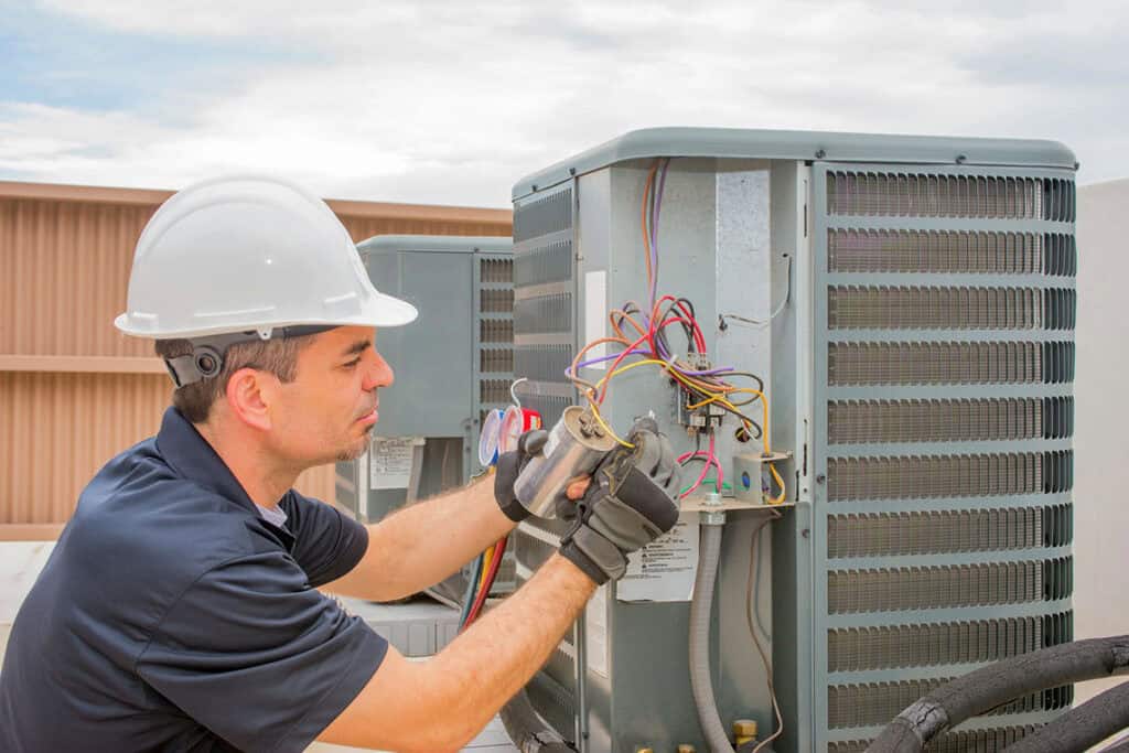 A technician in a hard hat and gloves performing maintenance on an outdoor HVAC unit, inspecting electrical connections and hoses.