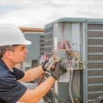 A technician in a hard hat and gloves performing maintenance on an outdoor HVAC unit, inspecting electrical connections and hoses.
