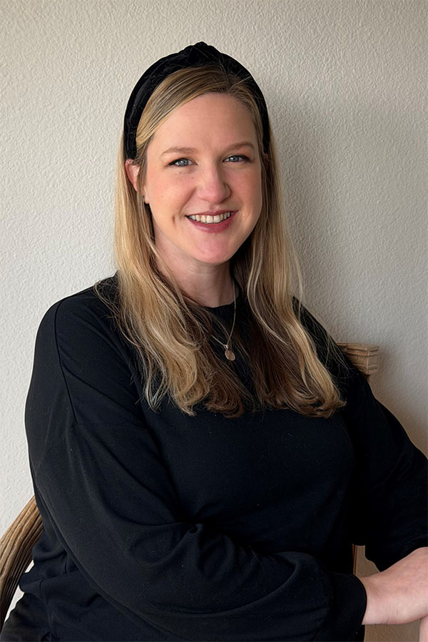 A woman with long blonde hair and a black headband smiles while sitting against a light-colored wall, wearing a black long-sleeve shirt.