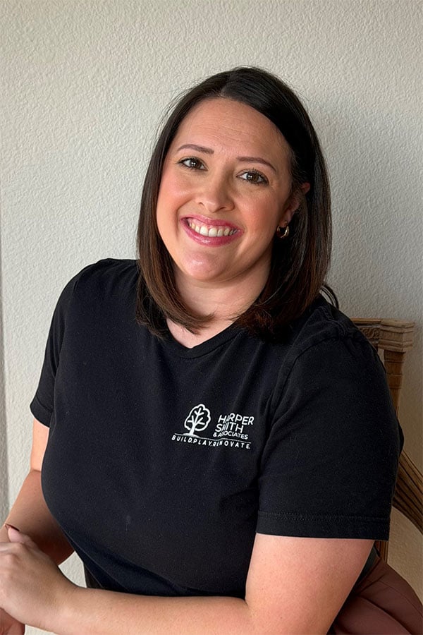 A woman with shoulder-length dark hair smiles while wearing a black t-shirt with a logo, seated against a light-colored wall.
