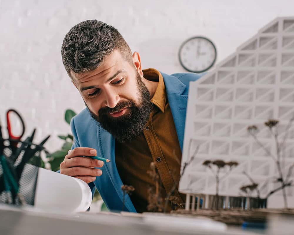 A man in a blue blazer and brown shirt intently examines a architectural model on his desk, holding a green pencil. Scissors and other drawing tools are visible in the background, along with a clock on the wall.