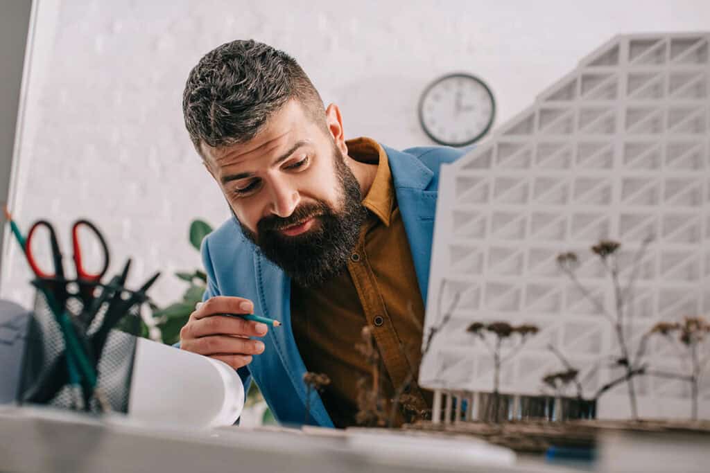 A man in a blue blazer and brown shirt intently examines a architectural model on his desk, holding a green pencil. Scissors and other drawing tools are visible in the background, along with a clock on the wall.