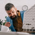 A man in a blue blazer and brown shirt intently examines a architectural model on his desk, holding a green pencil. Scissors and other drawing tools are visible in the background, along with a clock on the wall.